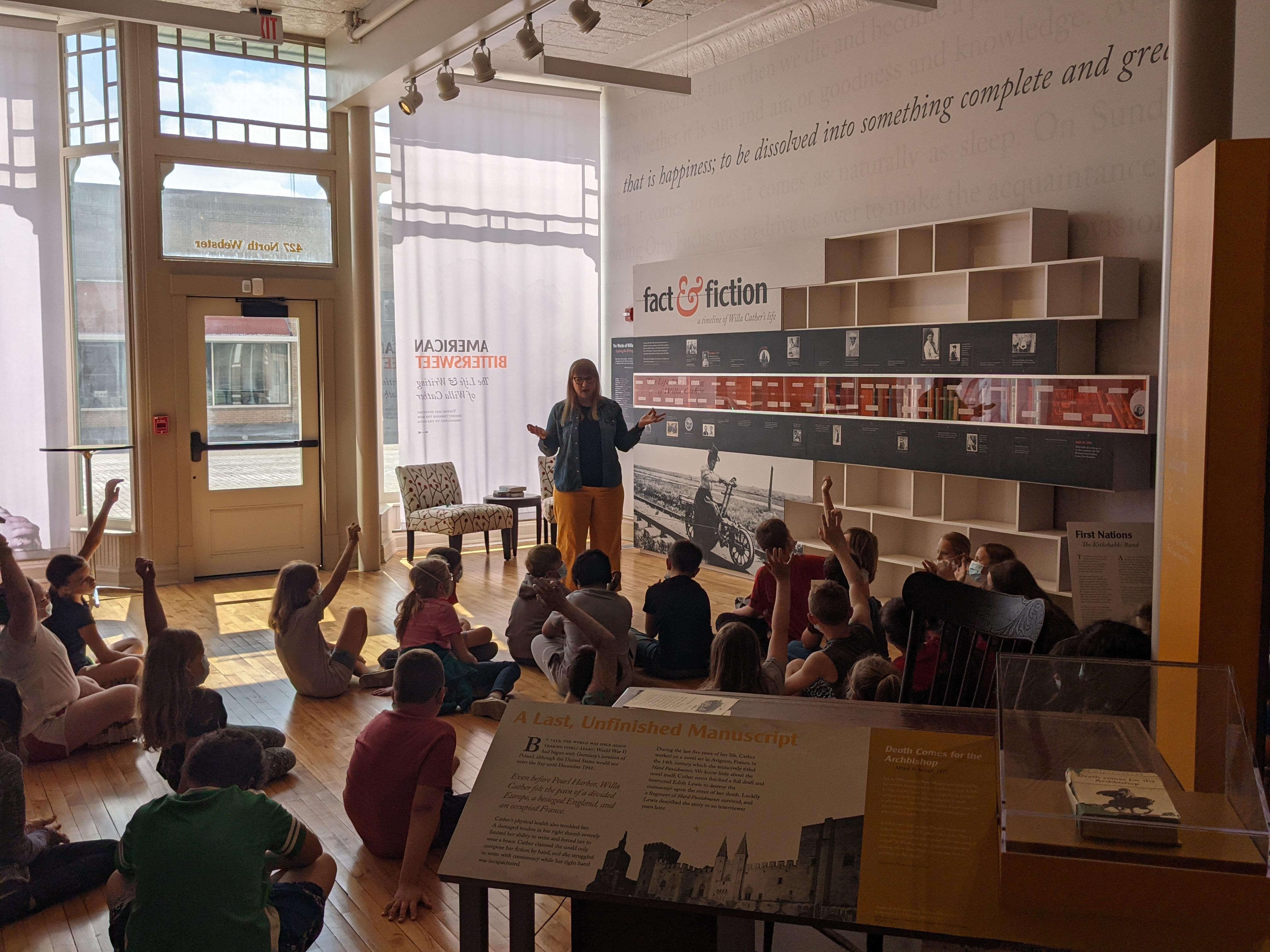 Education Director, Rachel Olson, speaks to a group of students sitting in front of a display in museum.